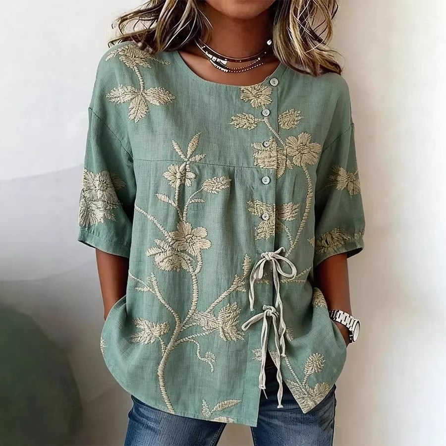Sage green blouse with cream floral embroidery, button-down front, and tie-detail, featuring short sleeves.