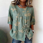 Sage green blouse with cream floral embroidery, button-down front, and tie-detail, featuring short sleeves.