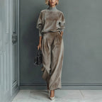 A model wears a taupe velour long-sleeve high-neck top and matching wide-leg pleated trousers with heels and a bag.