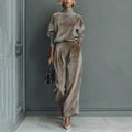 A woman wears a taupe velour high-neck long-sleeve top and matching pleated wide-leg pants against a gray wall.
