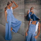 A woman wears a light-wash denim overall jumpsuit with a wide-leg silhouette over a white tank top.
