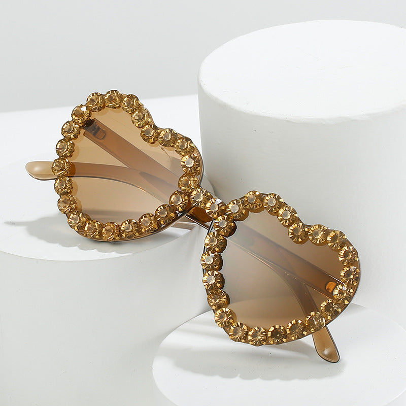 Heart-shaped sunglasses with brown tinted lenses and a gold crystal-encrusted rim, displayed on white pedestals.