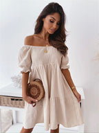 Woman wearing a beige, off-the-shoulder, smocked mini dress with puff sleeves, holding a round woven straw bag.
