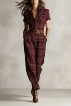 A model wears a burgundy plaid short-sleeved jumpsuit with a button-front, chest pockets, and a brown leather belt.