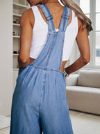 A woman wearing a white racerback tank top underneath light-wash blue denim overalls.