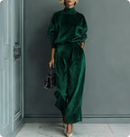 A person wears a dark green, velour high-neck long-sleeve top and matching wide-leg pants, holding a black handbag.