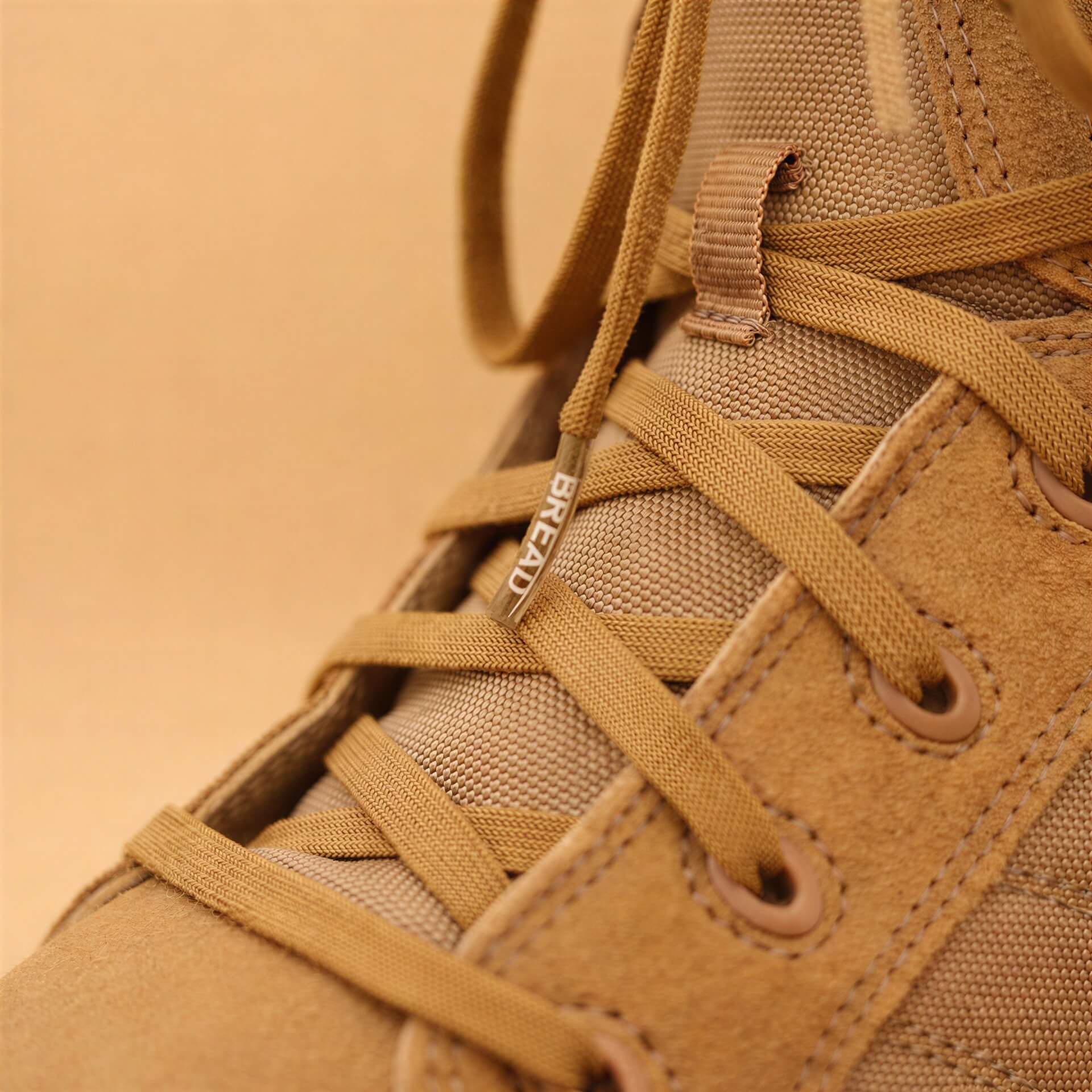 Close-up of a tan suede and canvas lace-up boot with a metallic aglet engraved with the word "BREAD."
