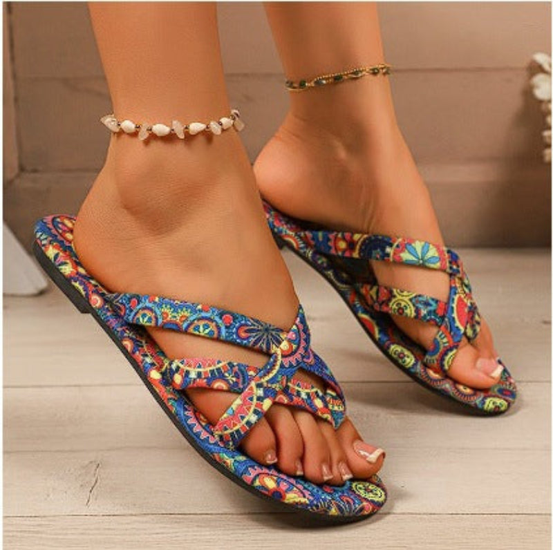 Flat sandals with vibrant blue, yellow, and red bohemian pattern, featuring crossed straps and a rounded toe.
