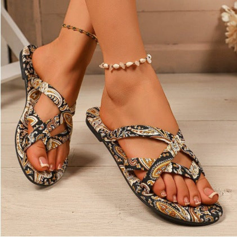Flat sandals with a black, white, and mustard yellow paisley print, featuring crisscross straps and a toe post.