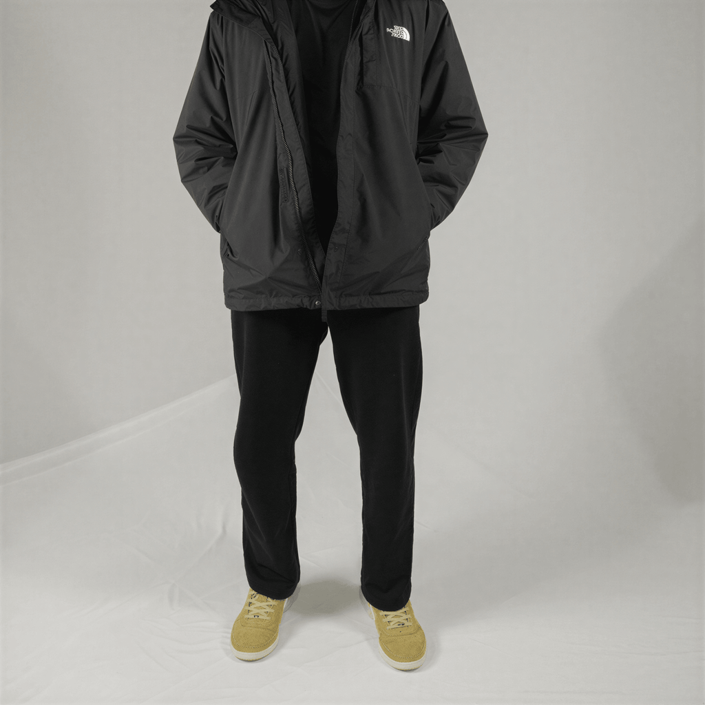 A person wears a black North Face hooded windbreaker, black pants, and tan suede sneakers against a grey background.