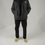A person wears a black North Face hooded windbreaker, black pants, and tan suede sneakers against a grey background.