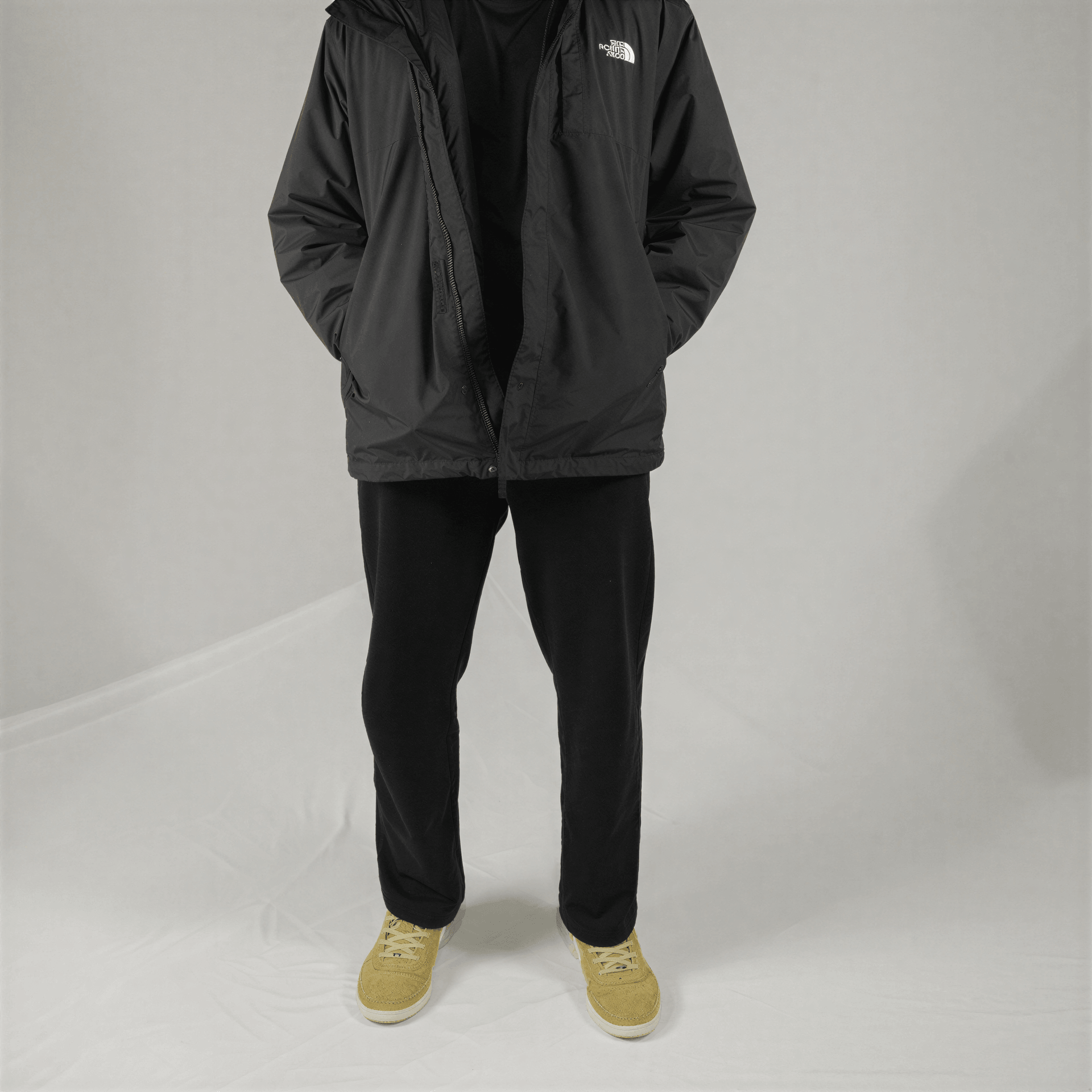 A person wears a black North Face hooded windbreaker, black pants, and tan suede sneakers against a grey background.