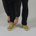 A person wears black trousers and mustard yellow suede sneakers with a small blue cross on the tongue.