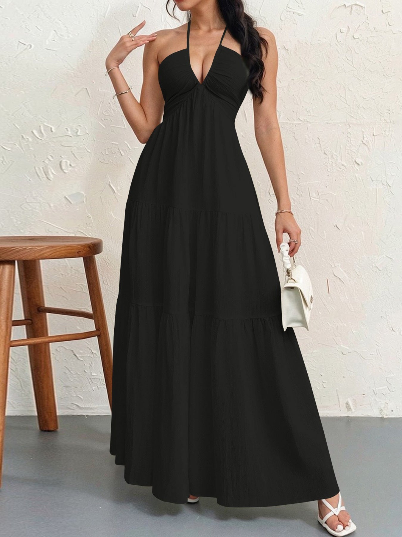 A woman wears a black, sleeveless halter maxi dress with a tiered skirt, holding a small white handbag.