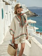 A woman in a cream linen poncho and shorts with red and green striped trim, paired with a matching fringe scarf.