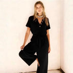 Model wearing a black short-sleeve jumpsuit with a button-front V-neck, tie waist, and wide legs.