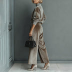 A woman models a taupe velour high-neck long-sleeve top and matching wide-leg trousers, holding a black quilted handbag.