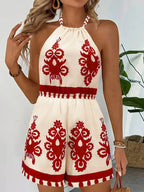 Cream halter romper with a red folk-inspired floral print and a defined, tassel-trimmed waist.