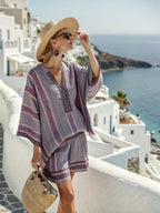 A woman wears a grey and purple striped tunic and shorts set with embroidered trim, a straw hat, and woven bag.