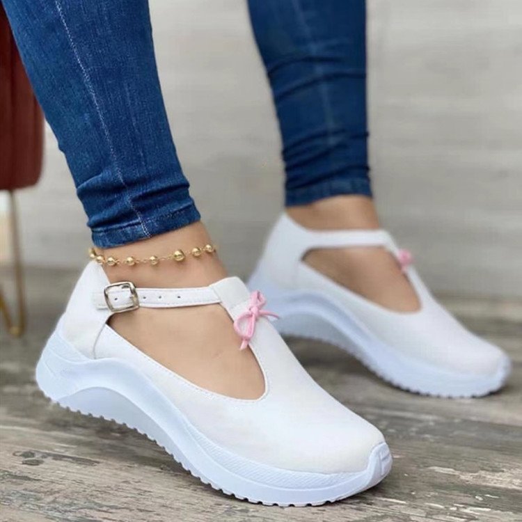 White T-strap sneakers with a side buckle, pink bow accent, and thick platform sole, worn with blue jeans.